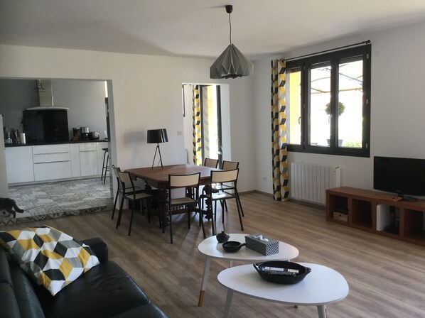 En moderne stue med spiseplads, et køkken i baggrunden og et stort vindue med gardiner.