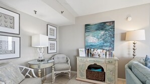 Un salon avec une table ronde, deux chaises, un canapé, un meuble avec un panier et un tableau accroché au mur.