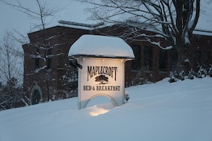 Letrero cubierto de nieve para el Bed & Breakfast Maplecroft, con un edificio de ladrillo al fondo.