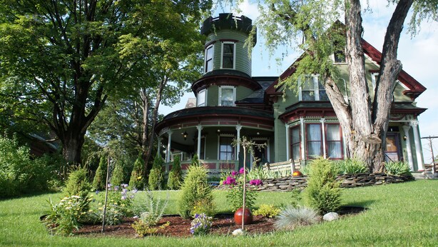 Una casa grande e histórica con torre y porche envolvente, rodeada de un jardín cuidado.