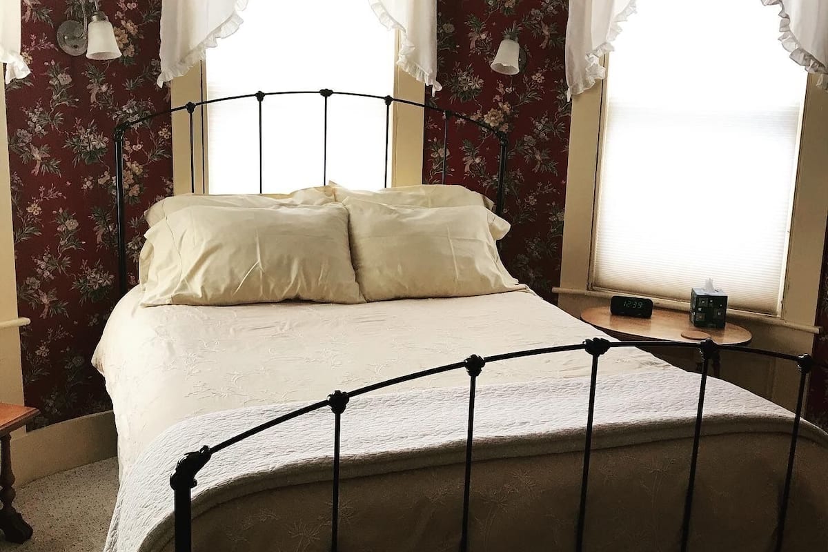 A bedroom with a metal bed frame, floral wallpaper, and two windows with white curtains.