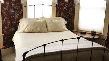 Quarto com estrutura de cama de metal, papel de parede floral e duas janelas com cortinas brancas.
