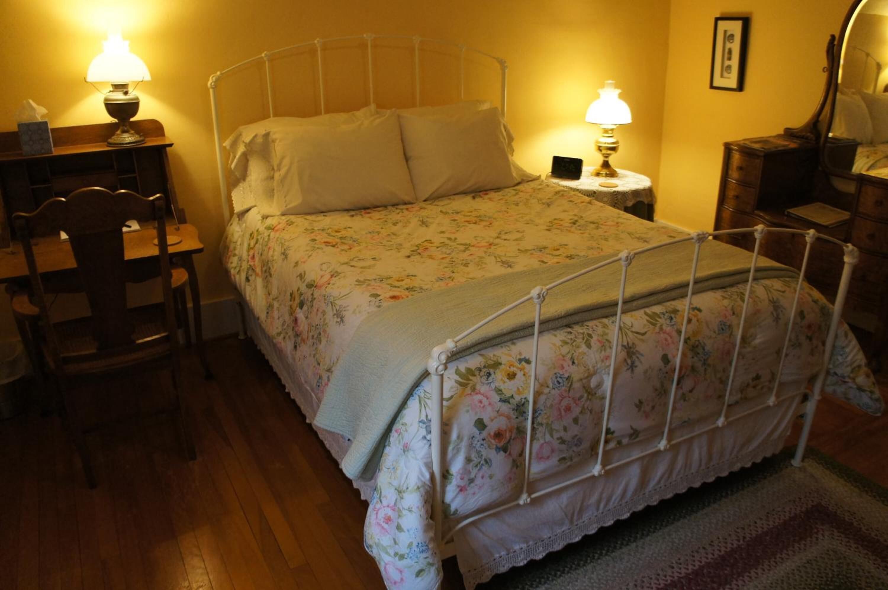 A bed with a metal frame, a wooden chair, a small bedside table with a lamp, and a wooden dresser.