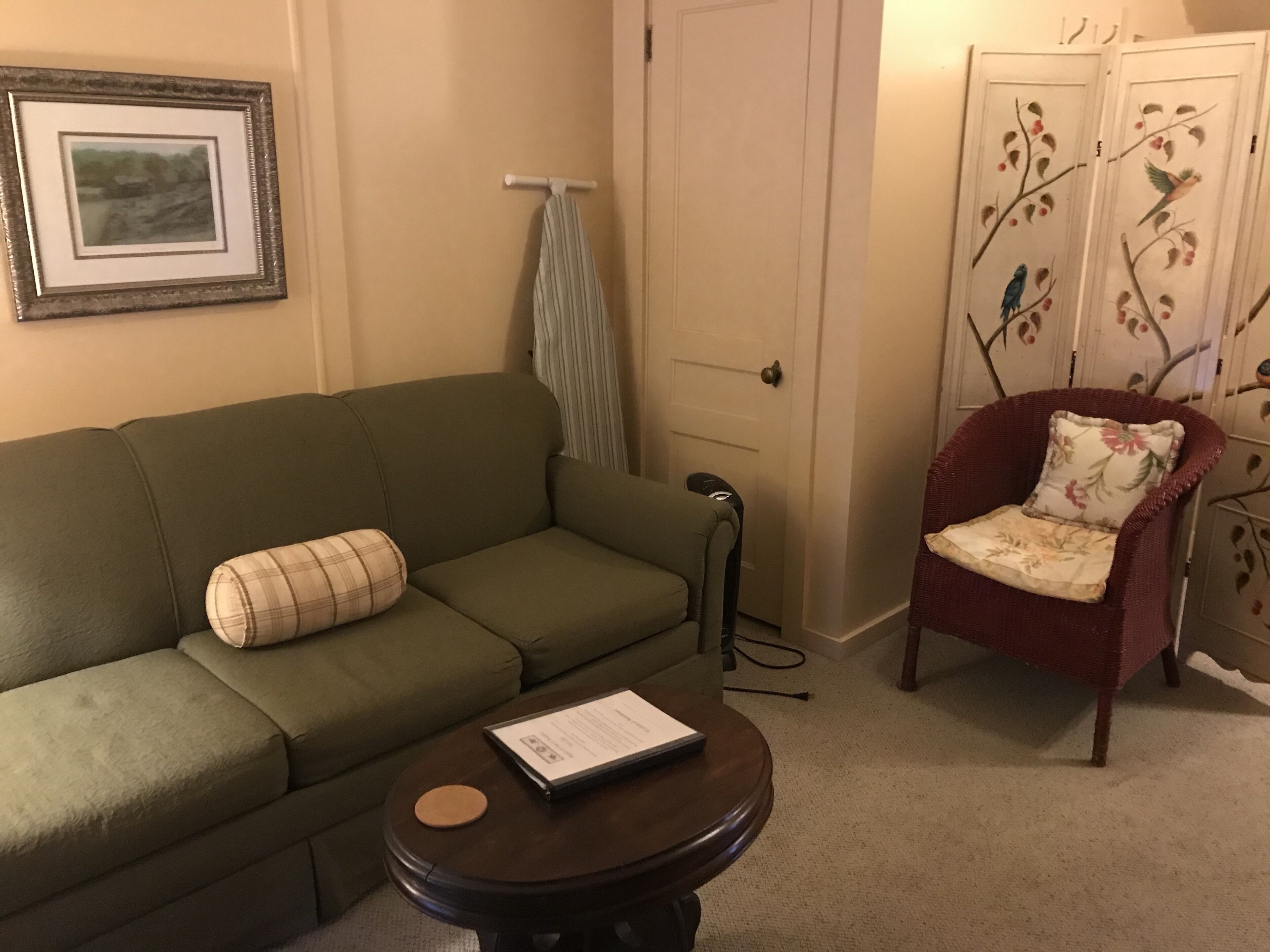 Una sala de estar con un sofá verde, una mesa redonda de centro con una computadora portátil, un sillón rojo con cojines estampados y un cuadro en la pared.