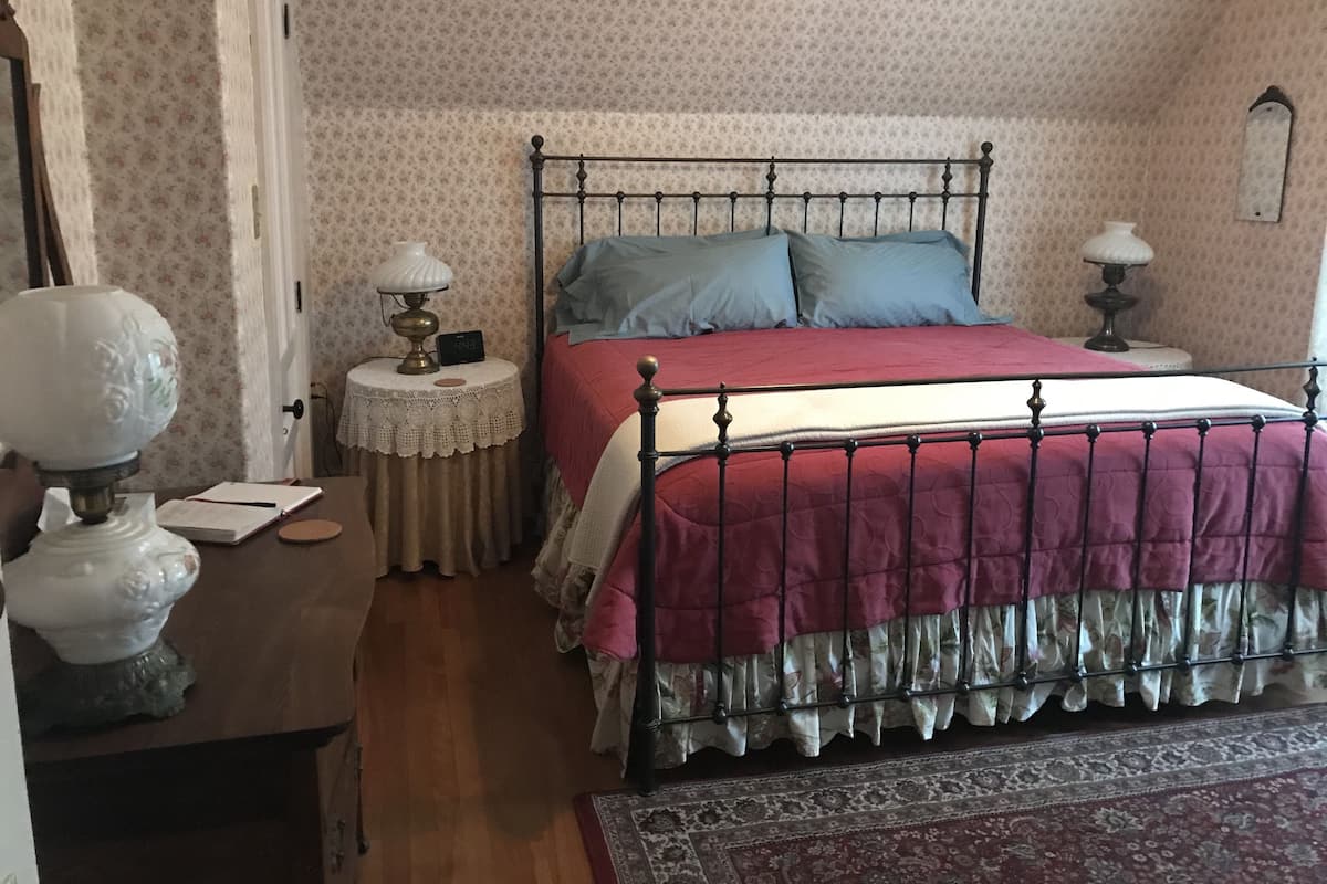 A bedroom with a metal bed frame, a red and white bedspread, two pillows, a nightstand with a lamp, a small table with a book, and a patterned rug.