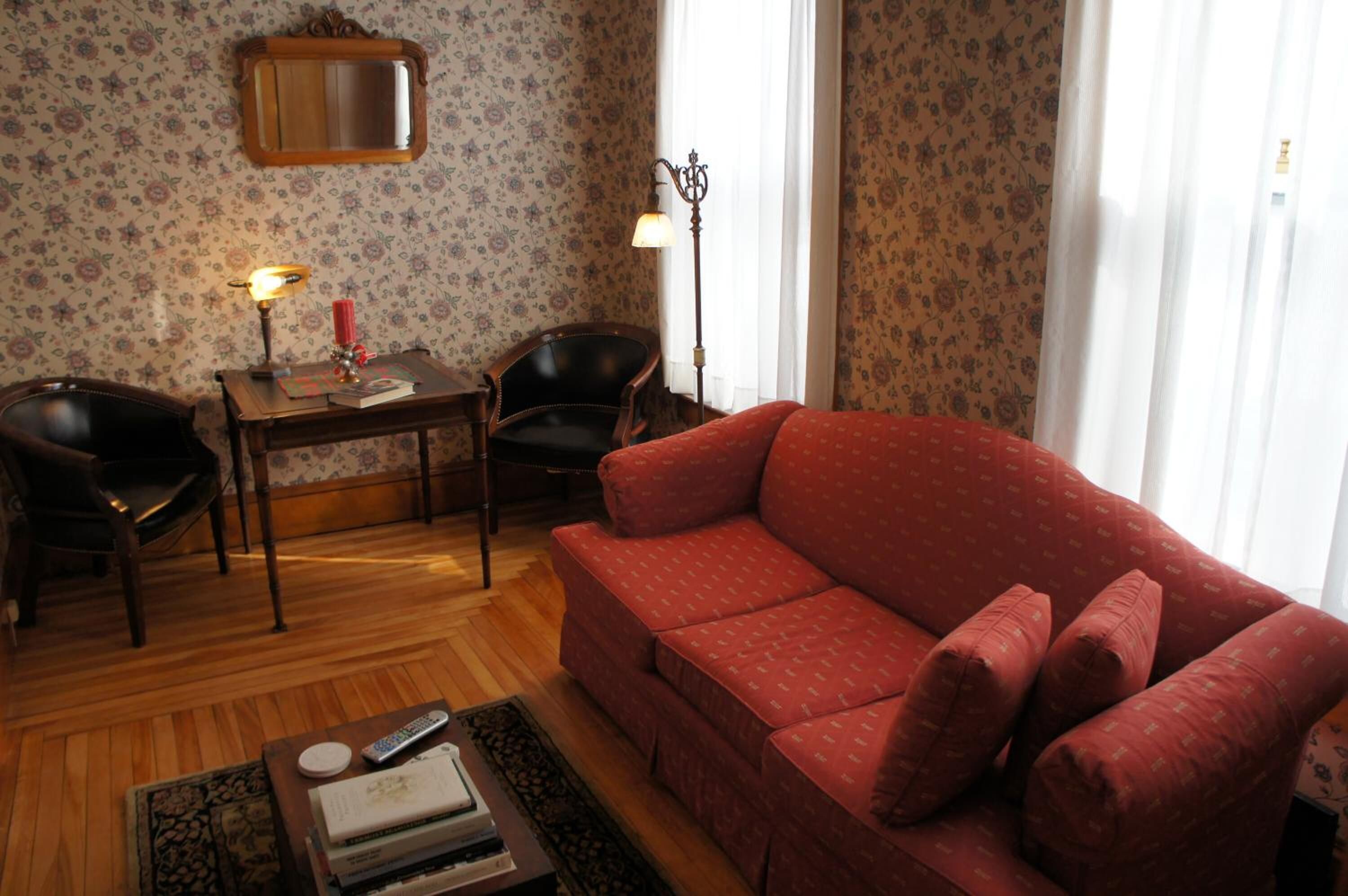 A living room with a red sofa, a wooden coffee table with books and a remote control, a small table with a lamp and a candle, and a chair.