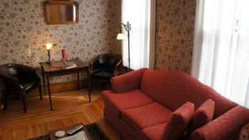 A living room with a red sofa, a wooden coffee table with books and a remote control, a small table with a lamp and a candle, and a chair.