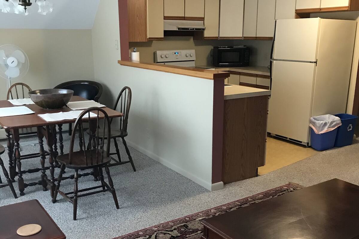 A compact living space with a dining area, kitchenette, and a small seating area with a patterned ottoman.