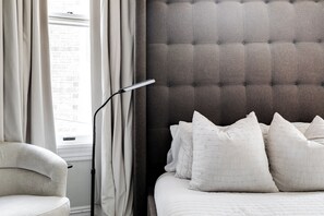 Un dormitorio con cabecera capitonada, cama con ropa de cama blanca, una silla y una lámpara de pie.