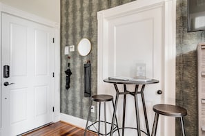 Una habitación con una puerta blanca, un espejo redondo, una pequeña mesa circular con dos taburetes y una pared con un patrón texturizado.