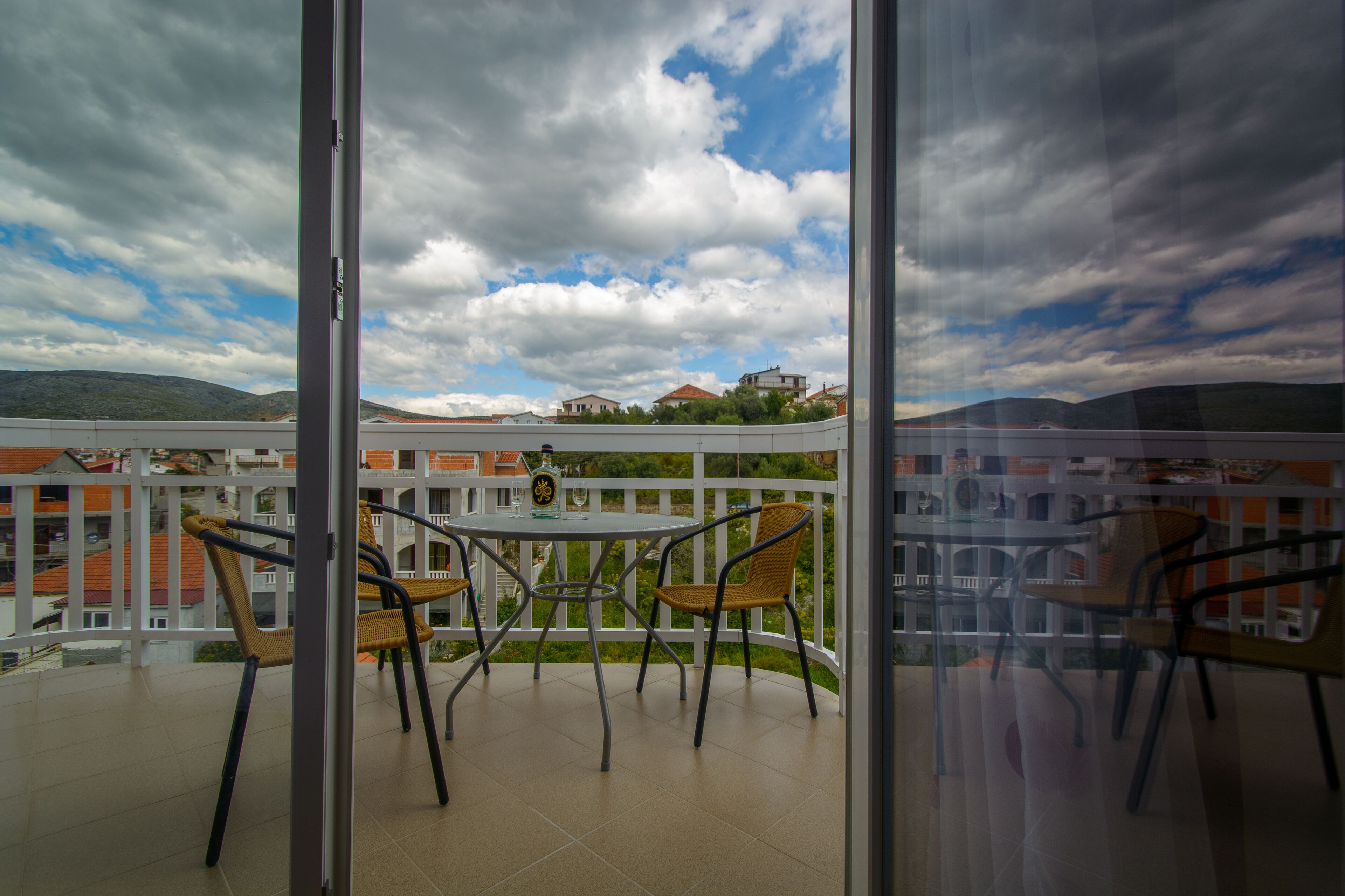 A balcony with a table and chairs, overlooking a residential area with houses and a mountainous backdrop.