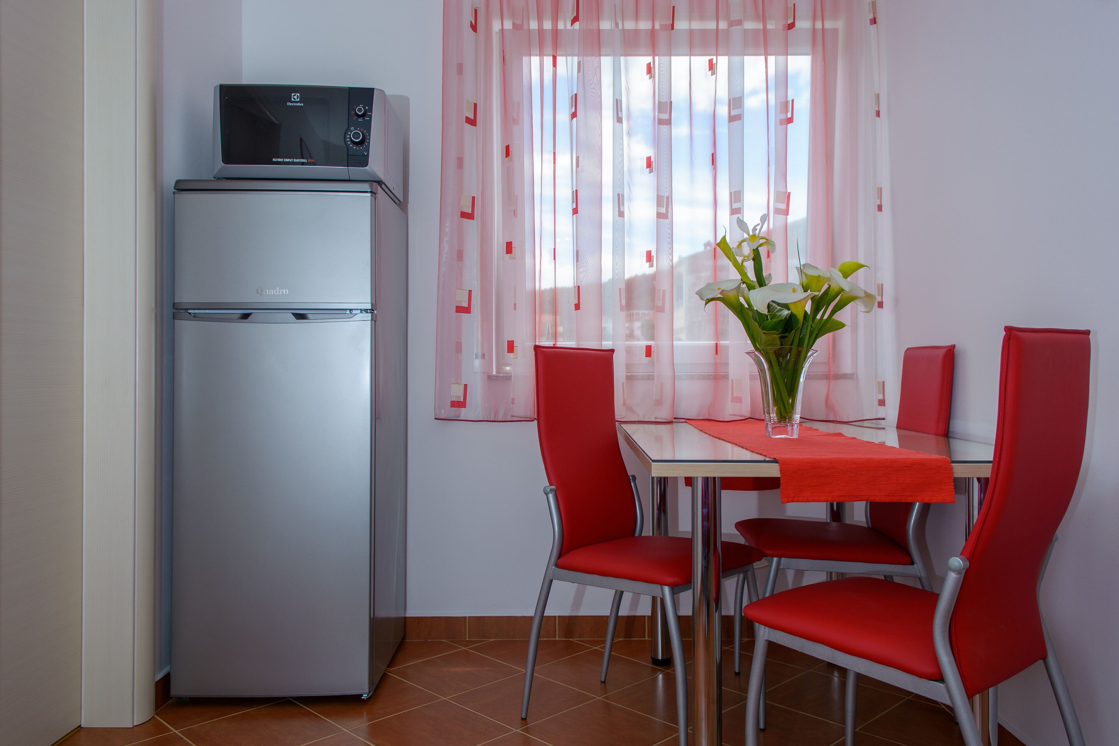 A compact kitchen with a refrigerator, a dining table with four red chairs, and a window with sheer curtains.