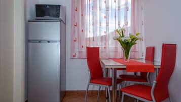 A compact kitchen with a refrigerator, a dining table with four red chairs, and a window with sheer curtains.