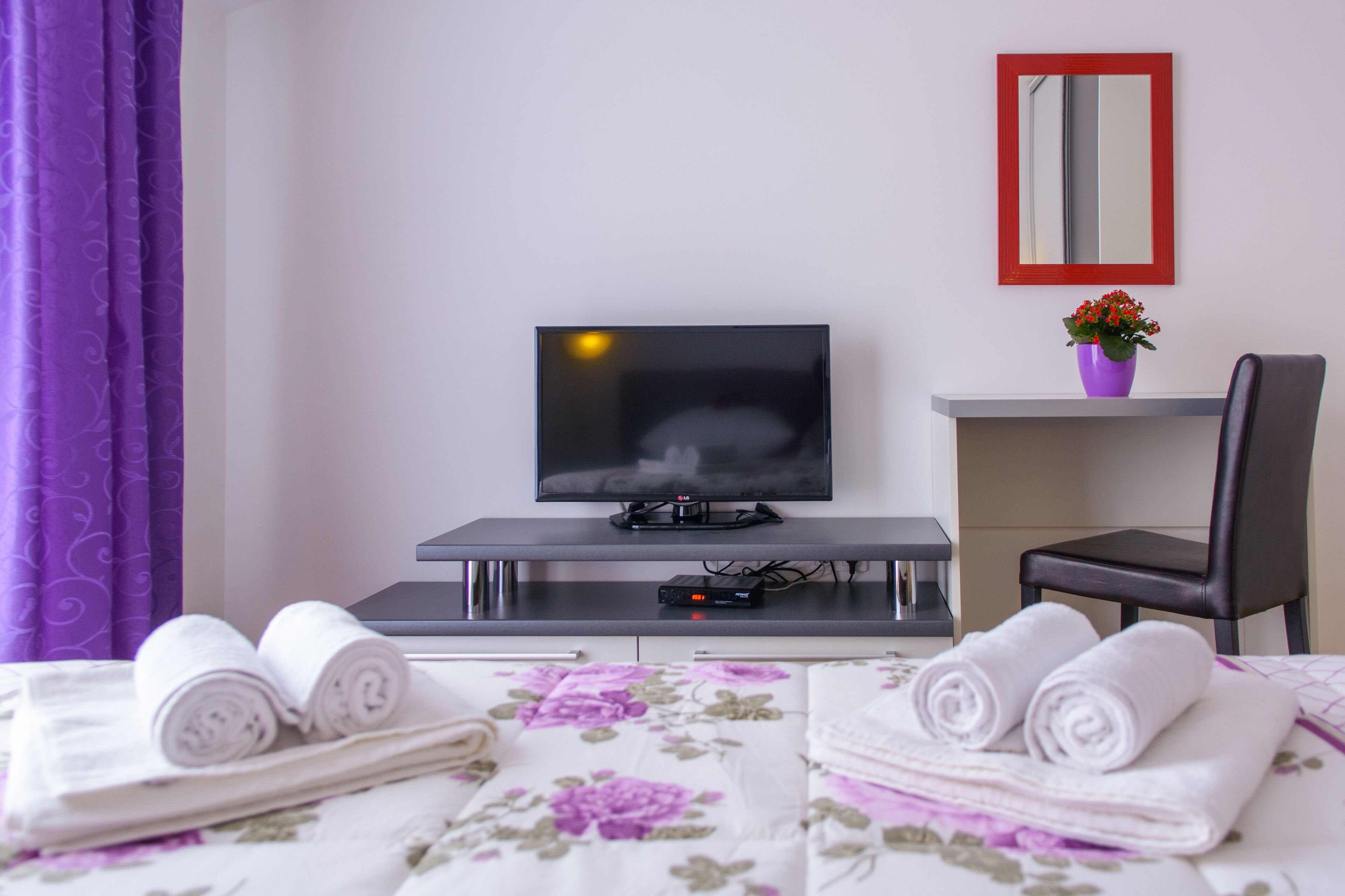 Quarto de hotel com uma cama, uma mesa de trabalho com televisão, uma cadeira e uma pequena mesa com um vaso de flores.