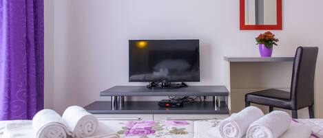 Quarto de hotel com uma cama, uma mesa de trabalho com televisão, uma cadeira e uma pequena mesa com um vaso de flores.