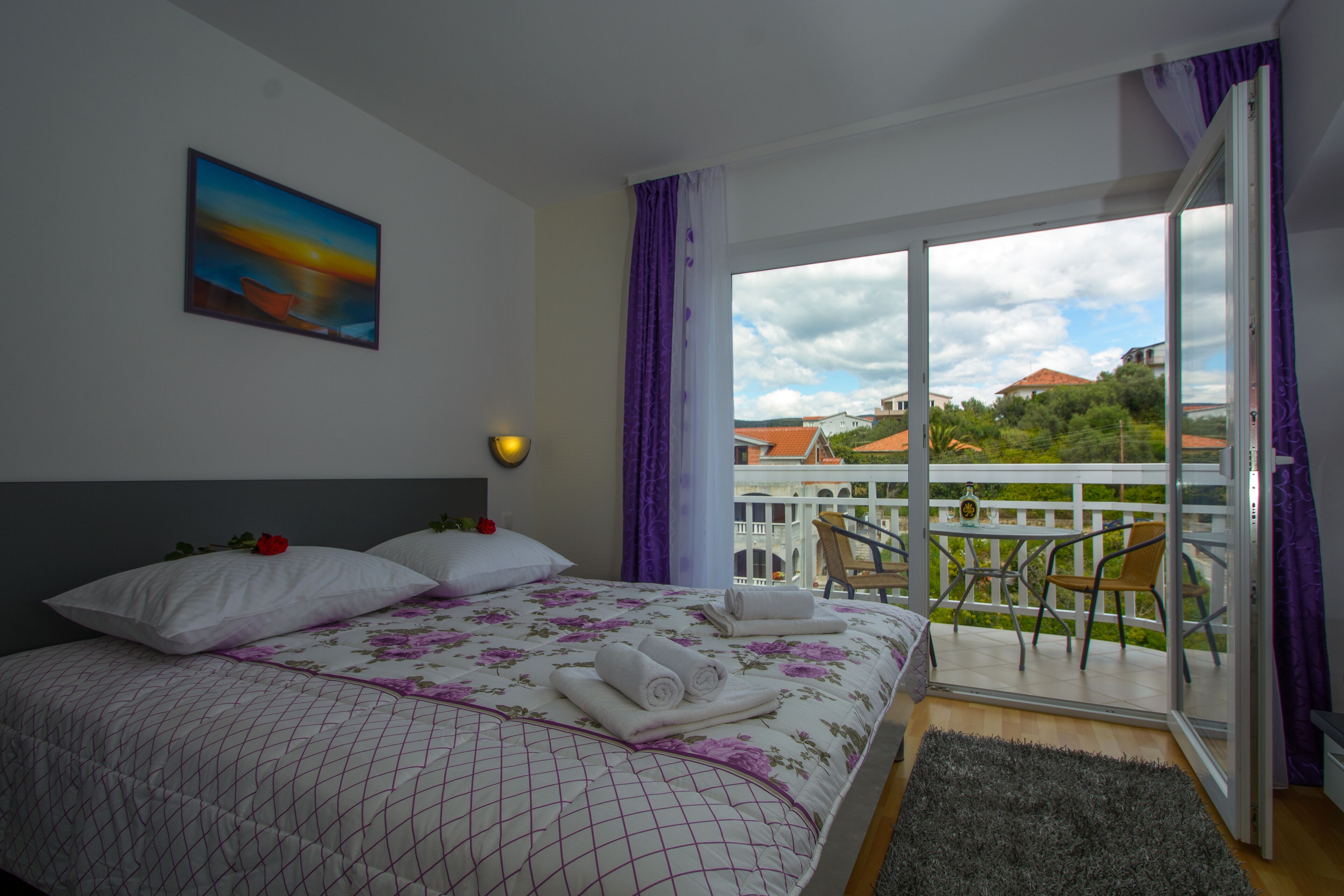 A hotel room with a bed, a painting, a balcony with chairs, and a view of buildings and greenery.