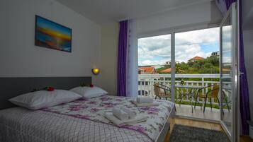 A hotel room with a bed, a painting, a balcony with chairs, and a view of buildings and greenery.