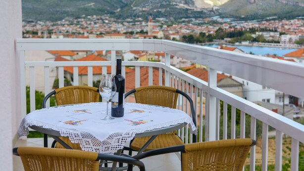 A balcony with wicker chairs and a table set for two, overlooking a town with red-roofed buildings and mountains in the distance. - Apartmani Mijo (Trogir)