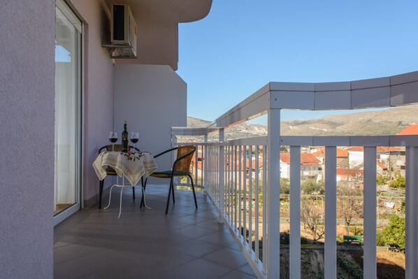 A balcony with a table and chairs, a view of a mountainous landscape, and a building with red roofs in the background. - Apartmani Mijo (Trogir)