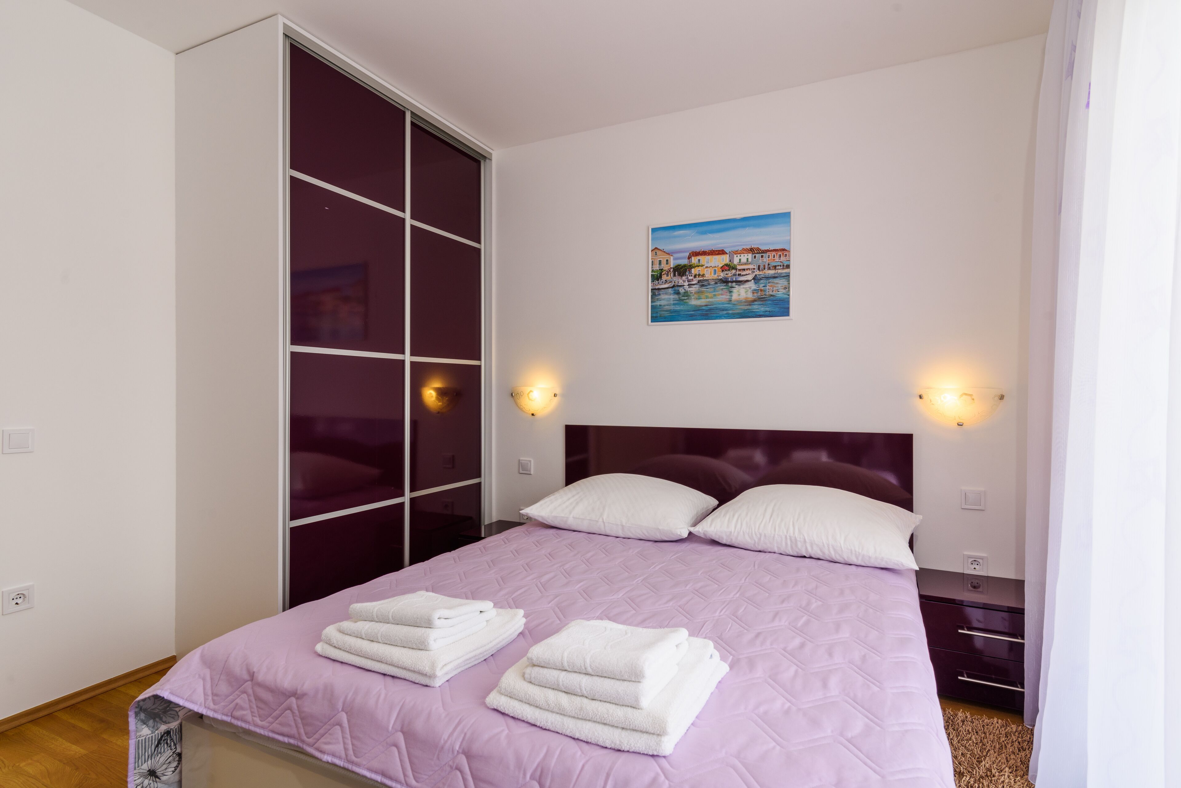 A neatly made bed with pillows and folded towels, a headboard, bedside tables, and a painting on the wall.