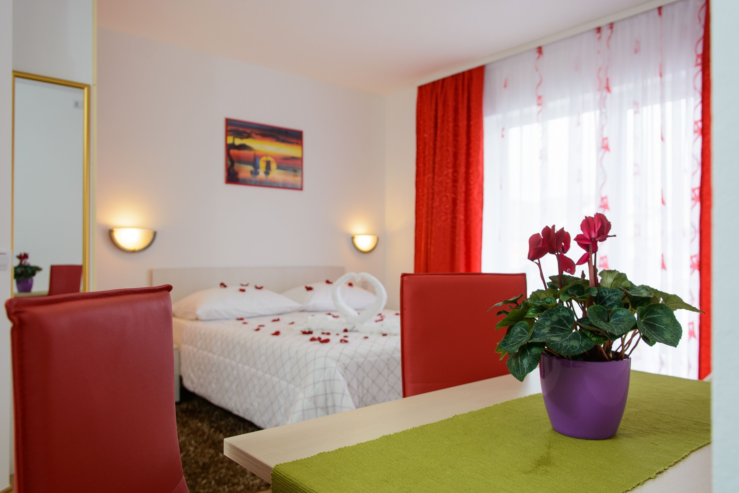 A hotel room with a bed, a desk, a chair, a potted plant, and a painting on the wall.