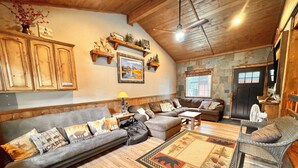 A cozy living room with a sectional sofa, a small table, and a wicker chair. There is a fireplace, wooden beams on the ceiling, and a stone wall. -  🌲20 Min to Yellowstone, High Speed Fiber, Hot tub, Fire Pit, Play Structure. (Island Park)