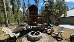 A wooden cabin with a deck, surrounded by trees. There is a fire pit with chairs and a table. -  🌲20 Min to Yellowstone, High Speed Fiber, Hot tub, Fire Pit, Play Structure. (Island Park)
