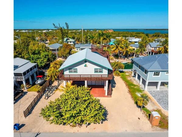 A single-story house with a covered porch, surrounded by a sandy driveway and a small garden. - Foster's Folly | Gorgeous canalfront pool home (Summerland Key)