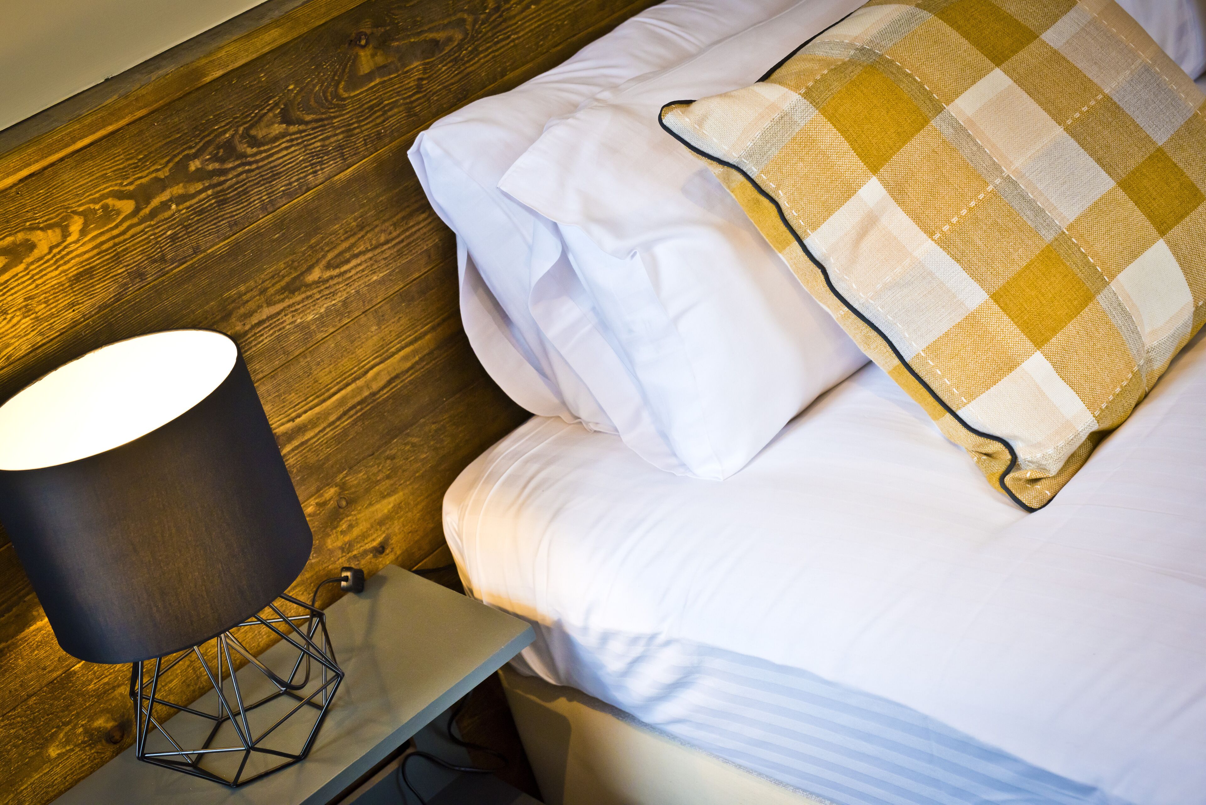 Una cama bien hecha con ropa blanca y una almohada de cuadros, una lámpara moderna sobre una mesita de noche de madera, y un suelo de madera.