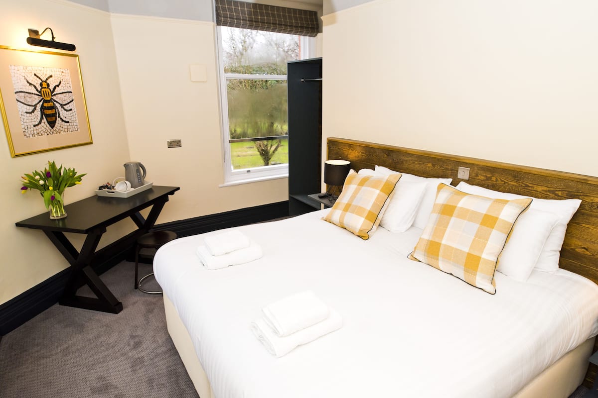 A neatly made bed with white linens and checkered pillows, a small desk with a kettle, a framed picture of a bee, and a window with a view of greenery.