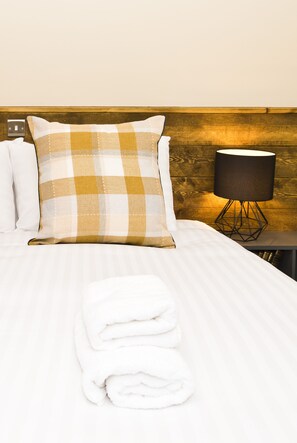 A neatly made bed with a checkered pillow, a folded white towel, and a lamp on a bedside table. - Bowling Green (Manchester)