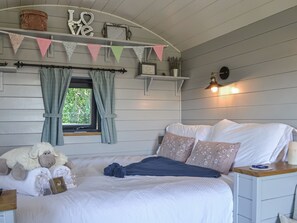 A bedroom with a bed, pillows, a bedside table, a window with curtains, a wall-mounted shelf with decorative items, and a wall-mounted light fixture. - 1 bedroom accommodation in Strensall, near York (Strensall, near York)
