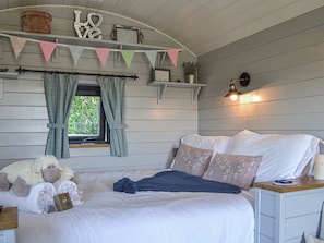 A bedroom with a bed, pillows, a bedside table, a window with curtains, a wall-mounted shelf with decorative items, and a wall-mounted light fixture.