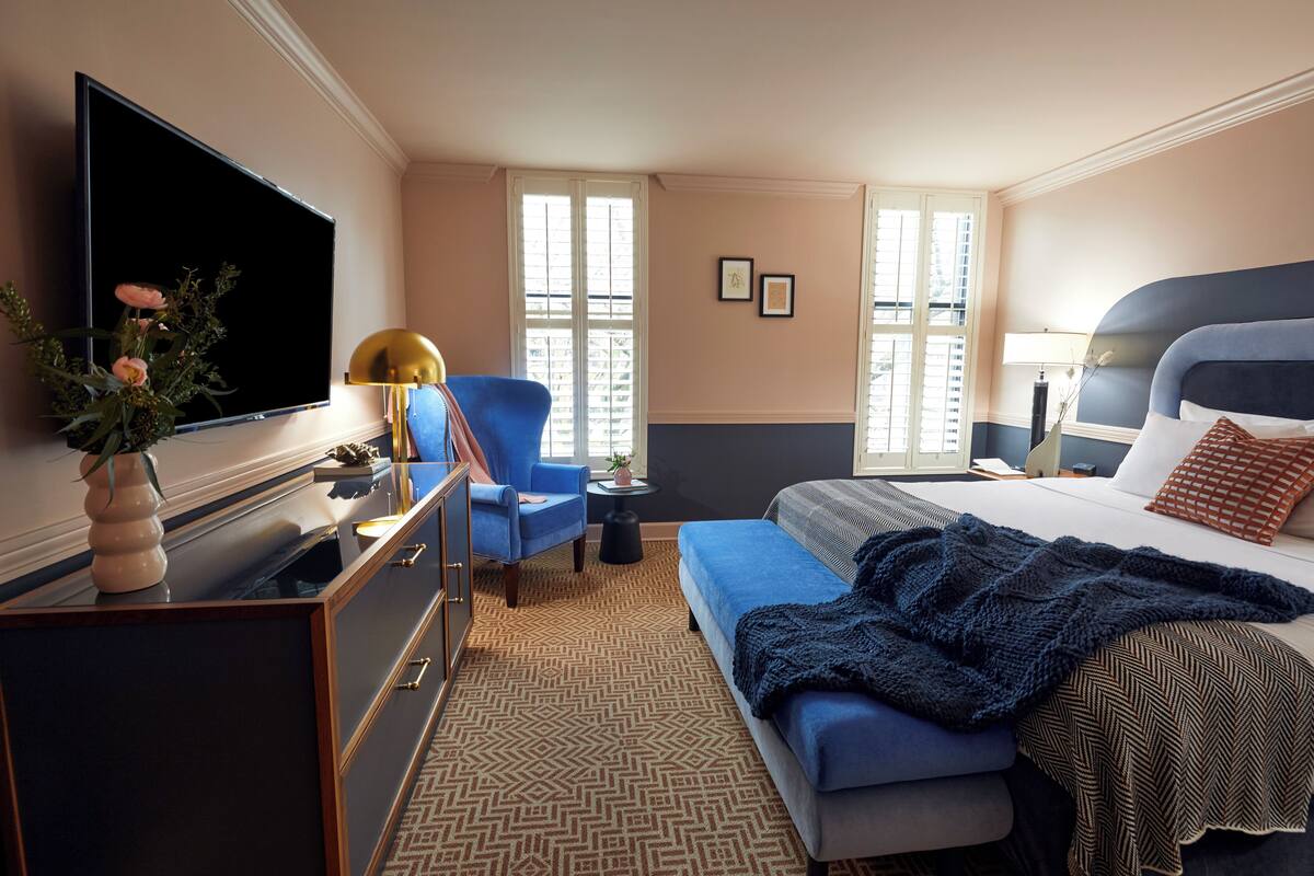 Habitación de hotel con cama, escritorio con televisor, silla, mesita pequeña y ventana con persianas.