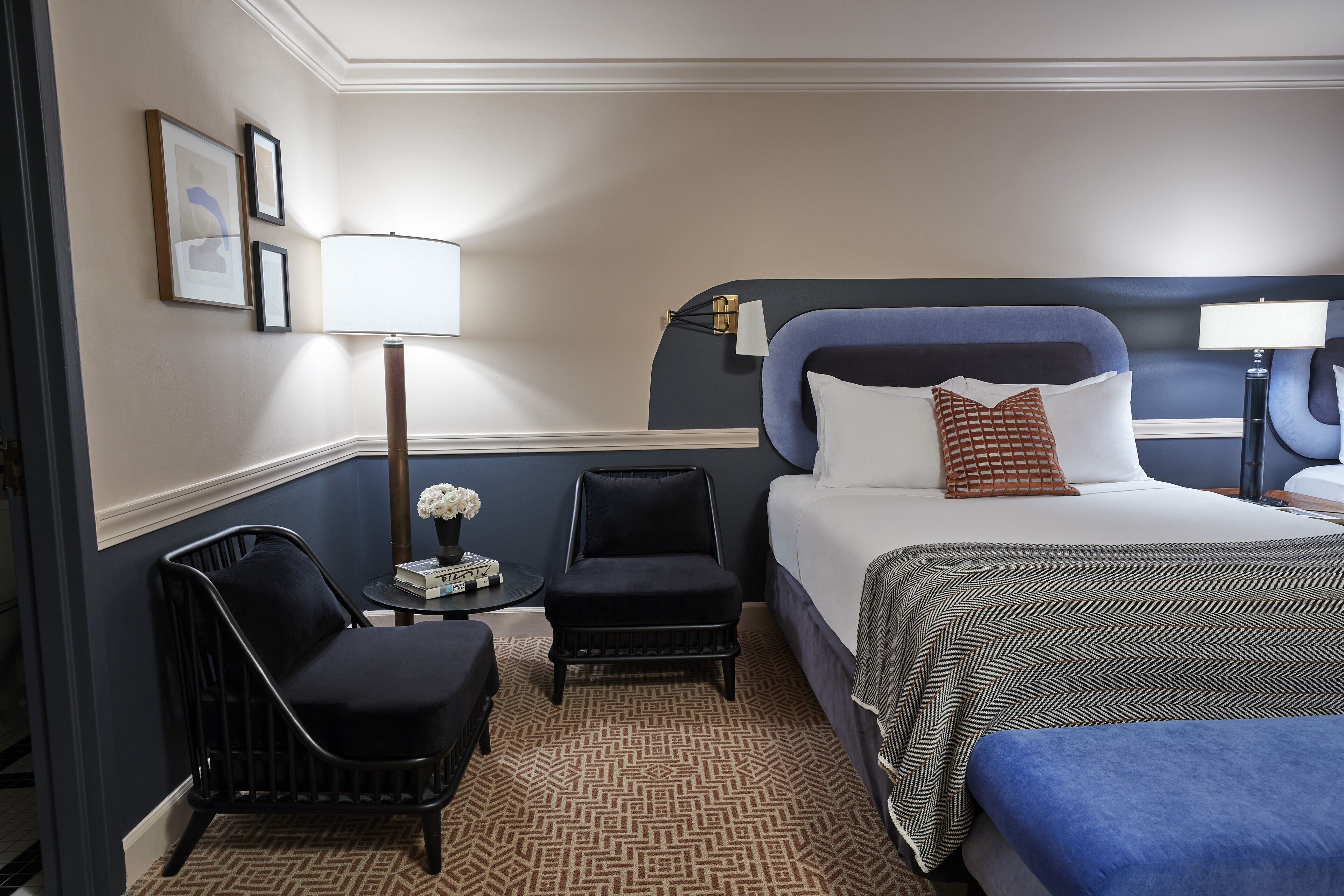 A hotel room with a bed, a chair, a small table with a lamp, and framed pictures on the wall.