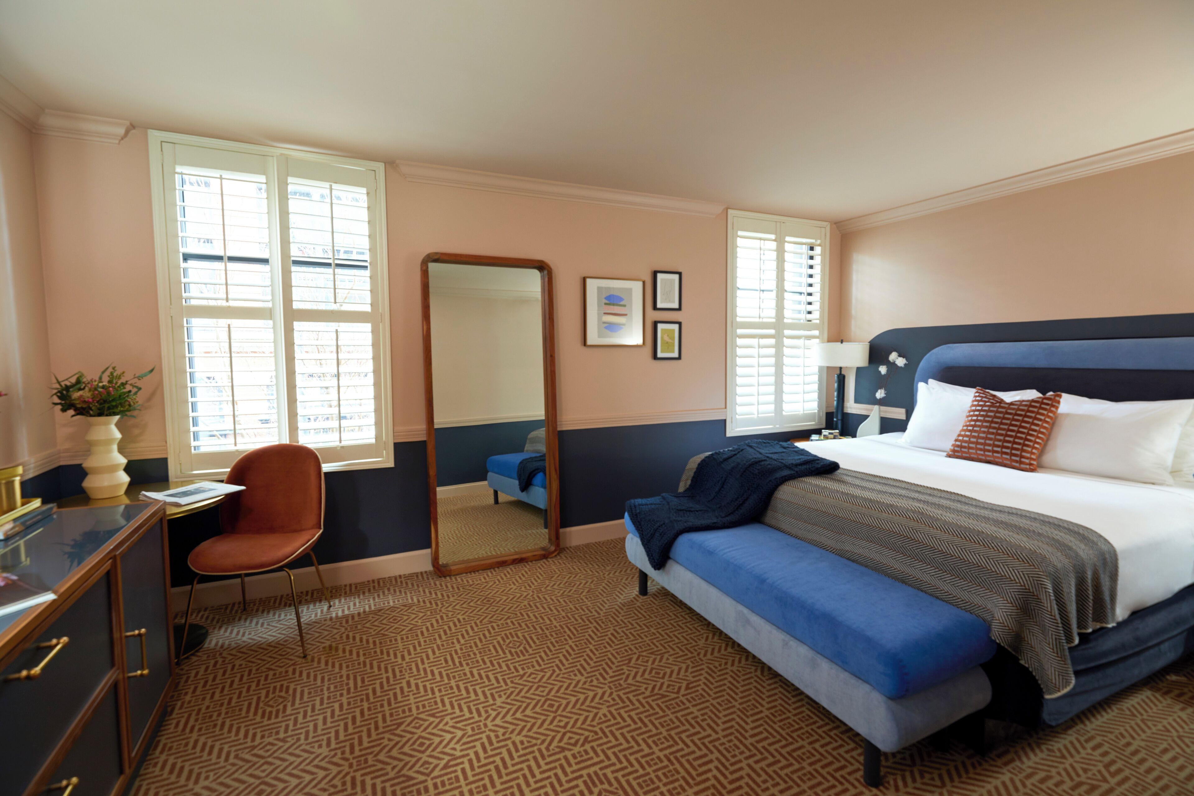 A hotel room with a bed, a desk, a chair, a mirror, and a window with blinds.
