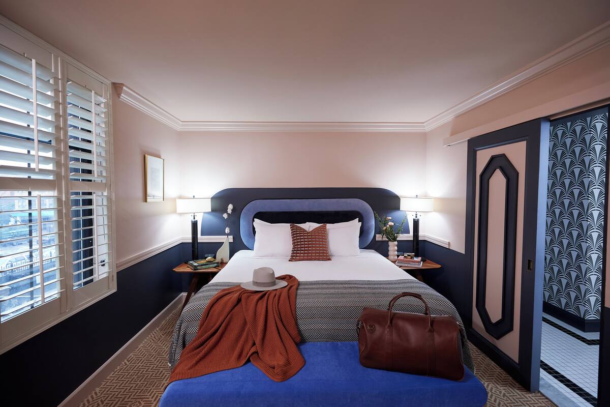 Habitación de hotel ordenada con una cama grande, una mesita de noche, persianas en una ventana y un papel tapiz estampado.