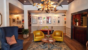Un salon spacieux avec un plafond haut, un grand lustre et un plancher en bois. On y trouve deux fauteuils jaunes, un fauteuil bleu et une table basse sur laquelle est posé un vase de fleurs.