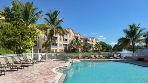 A pool area with lounge chairs and palm trees, surrounded by a building with balconies. - Bella Lago 241 by Distinctive Beach Rentals (Fort Myers Beach)