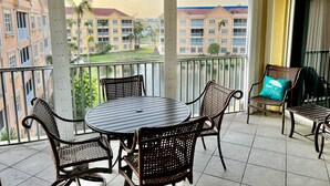 A balcony with a round table and wicker chairs, overlooking a residential area with buildings and a body of water. - Bella Lago 241 by Distinctive Beach Rentals (Fort Myers Beach)