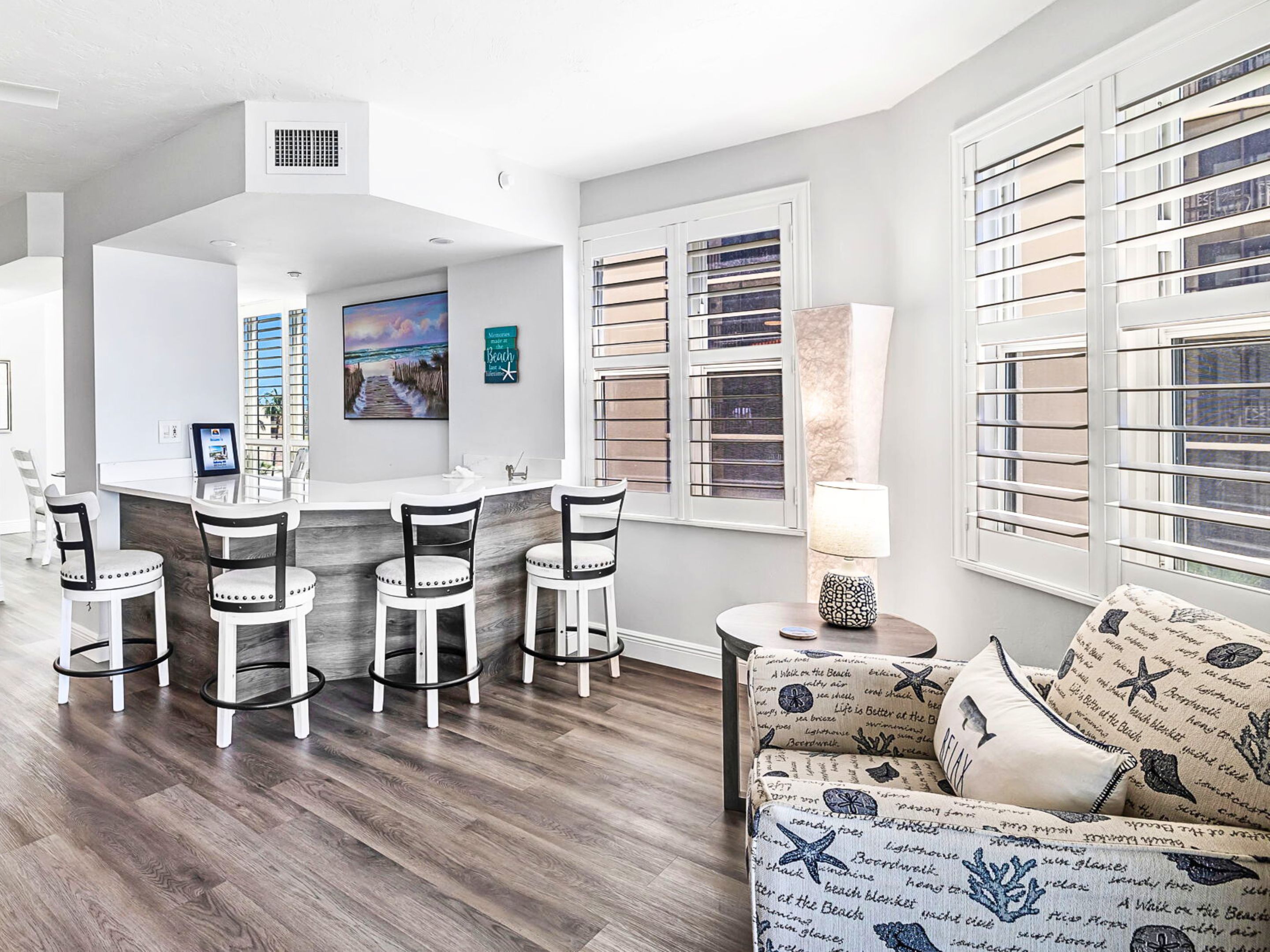 A modern living room with a kitchen area, featuring a bar with stools, a sofa with patterned cushions, and large windows with blinds.