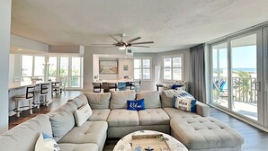 A spacious living room with a large sectional sofa, a dining area with a kitchen in the background, and a balcony with a view of the beach. - Gullwing 405 by Distinctive Beach Rentals (Fort Myers Beach)