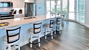A modern kitchen with white cabinetry, stainless steel appliances, and a large island with bar stools. - Gullwing 405 by Distinctive Beach Rentals (Fort Myers Beach)