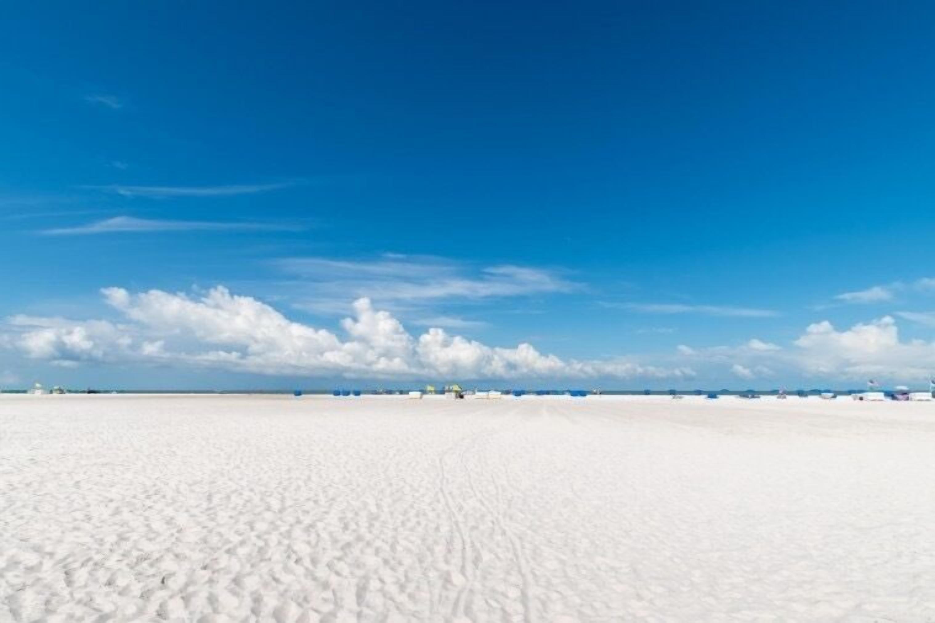 Una extensa playa de arena blanca con cielo azul despejado y algunas nubes dispersas.