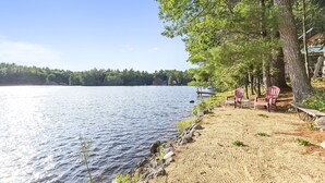 A lakeside view with a sandy beach, a row of Adirondack chairs, and a wooden deck.