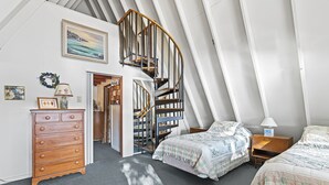 A bedroom with a wooden dresser, two beds with floral bedding, a spiral staircase, and a painting of a seascape.