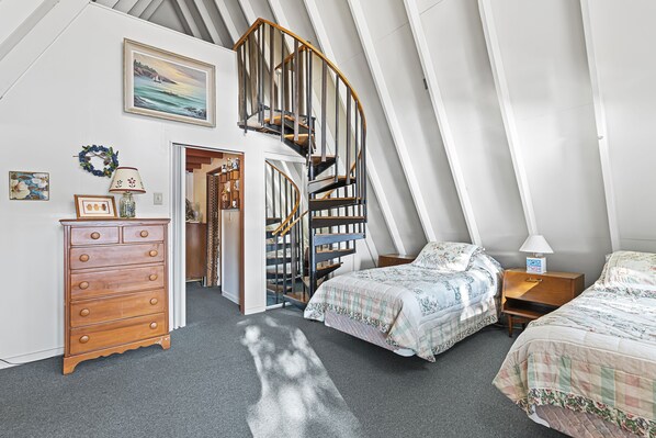 A bedroom with a wooden dresser, two beds with floral bedding, a spiral staircase, and a painting of a seascape.