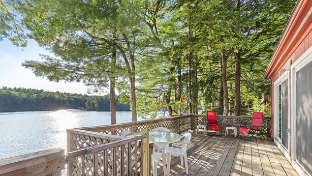 A wooden deck with a glass table and chairs, overlooking a lake with trees.
