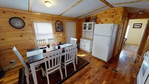 A wooden cabin interior with a dining table, chairs, a refrigerator, and a clock on the wall.
