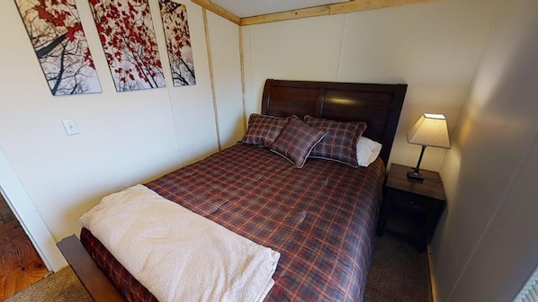 A single bed with a plaid bedspread and pillows, a bedside table with a lamp, and wall art depicting trees.